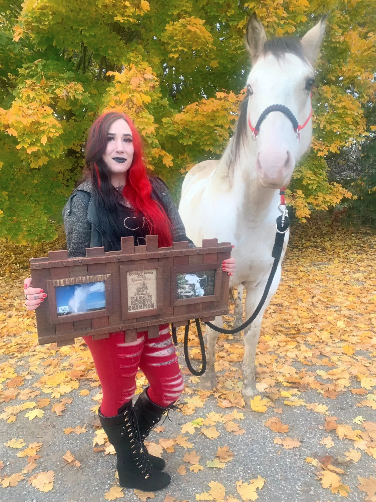 Jenna holding a frame of show photos beside her paint horse in autumn leaves