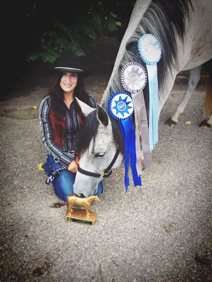 Jenna kneeling beside her grey horse with a row of blue show ribbons